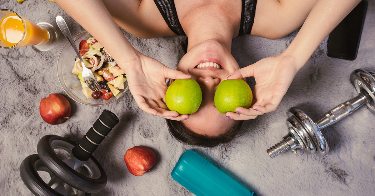 woman lying next to workout equipment and holding apples against her face
